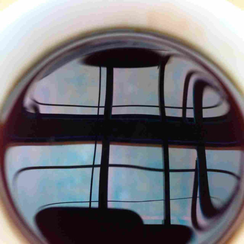 A circular liquid image surrounded by white. The white is a coffee mug lightly stained town from coffee. In the reflection from the coffee surface is a view above through a skylight. Black lines from window frames and wires crisscross. Beyond is a pinkish, blueish sky coloring sky and clouds.
