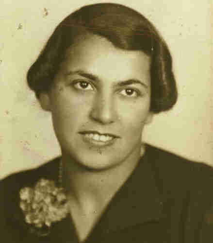 Portrait photograph of a young woman. She is smiling slightly, exposing her upper teeth. Her hair is neatly combed reaching her ears. She is wearing a dark jacket, with a flower on the left side of the frame. She is also wearing beads.