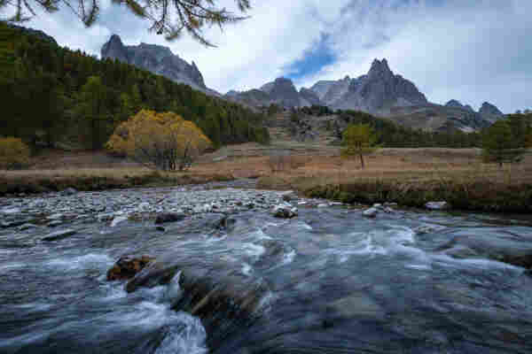 A mountainous landscape with a flowing river in the foreground, surrounded by autumn-colored trees and rugged mountain peaks in the background under a cloudy sky.