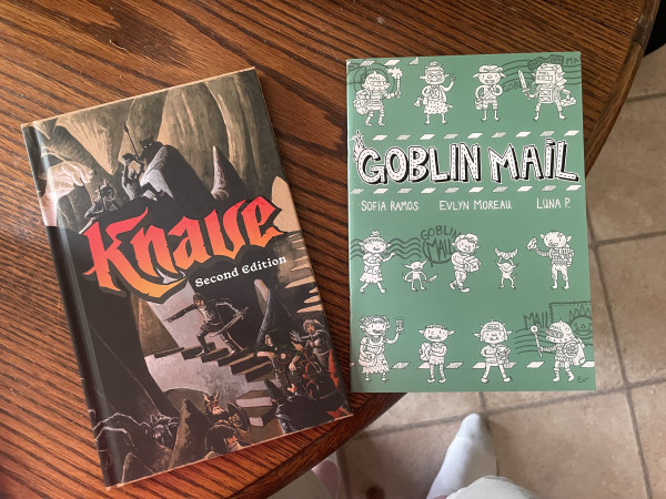 Two books on a wooden surface: "Knave: Second Edition" with a fantasy-themed cover featuring a dungeon scene, and "Goblin Mail" with a green cover and cartoonish goblin illustrations.