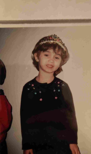 Erika as a child smiling while wearing a toy crown. She has a black dress with needs on it.