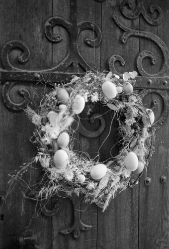 Black and white portrait format photo showing a garland hanging on an old door with ornate ironwork. The garland bizarrely features almost a dozen eggs!