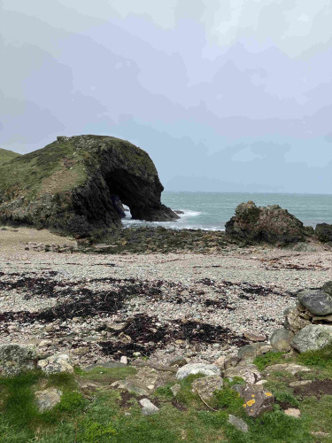 Rocky beach with a large arched sea cave in a hill, overcast sky, and choppy sea waters on the Anglesey coast.