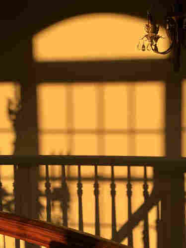 The light and shadow from a window and a railing on a beige wall just before sunset. It's actually 3 windows side-by-side with a fourth arched window above, but only the edges of the two side window are visible. Part of the railing is visible.
