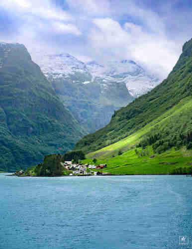 Color photo taken from the waters of a fjord looking towards a small village on the shore at the base of a steep mountain slope. In the background are more mountains rising from the fjord with snow on their peaks. Above is a partly cloudy sky. 