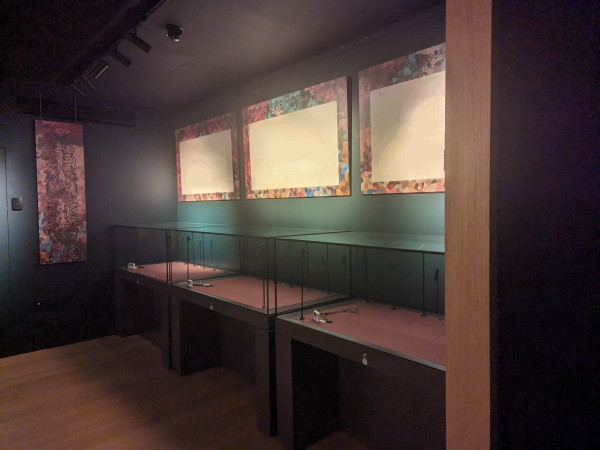 Green walled exhibition room with wooden floors. Overhead lights shining on three (redacted) panels with multi-coloured borders.
Three empty display cases each with small datalogger in the front corner.
