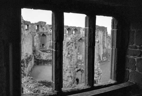 Looking out through a stone window, with two vertical stone divides, onto buildings in the ruins of Kenilworth Castle. Black and white photo.