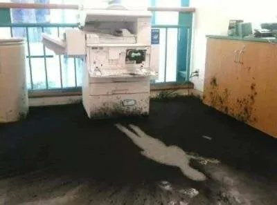 Still image. Copier that's spewed jet-black toner all over the carpeted floor within a six foot radius. There is a clean spot in the shape of a small human on the floor. 