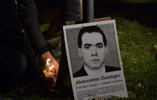 Ein schwarz-weiß-Plakat von Abdurrahim Özüdoğru. Es lehnt an einem Mast, daneben Hände, die Kerzen aufstellen.