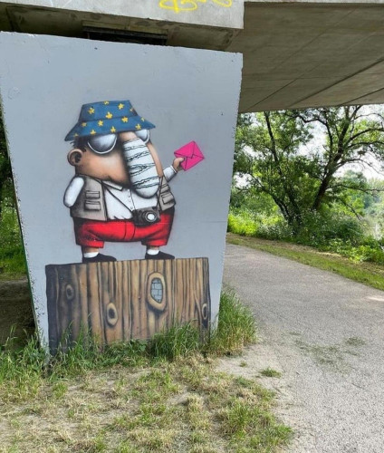 Streetartwall. The comic figure of a slightly damaged man with a large nose was sprayed/painted on the gray concrete wall under a bridge. The little man is wearing a blue hat with yellow stars (European flag), sunglasses as camouflage, short red trousers, beige sleeveless jacket, white T-shirt and a camera around his neck. His very long bandaged nose is a bit strange and unfortunately his arm without a hand also seems to be bandaged. He is standing on a tree stump and holding up a red election envelope. If he can go to the polls, so can the rest of the world, right? Title: "The people of tomorrow belong to us" (in french)