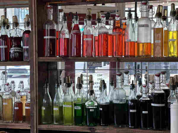 Glass bottles of colorful soda flavorings on shelves with mirrored background. On the top shelf is hues of red. Underneath is a shelf with hues of green and purple. Each has a small label naming its favorite. There is: strawberry, pink lemonade, tigers blood, guava, papaya, peach, orange, mango, pineapple seen on visible labels. Also: Carmel, honey, pumpkin spice, bubble gum, pomegranate, grenadine, lemon, banana, green apple, kiwi, mint, lime, green river, blueberry, grape, blackberry.