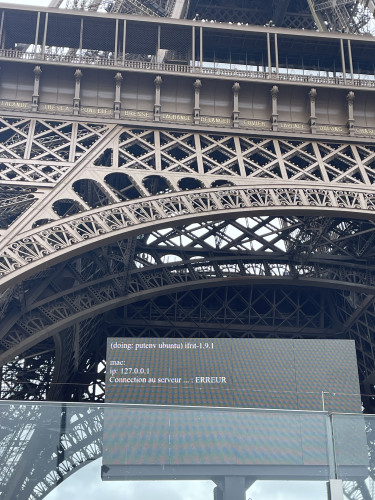 Photo of the Eiffel Tower, taken just below the tower. In the foreground there’s some sort of public info screen. It’s showing a Ubuntu shell that’s reporting a network problem.