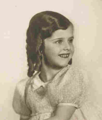 Photo of a young girl on a solid background. She can be seen from the waist up. The girl is wearing a dress with buff short sleeves. Her hair is neatly styled - the curls fall to the right side of her head and almost reach her shoulders. She is smiling tightly, exposing her upper teeth.