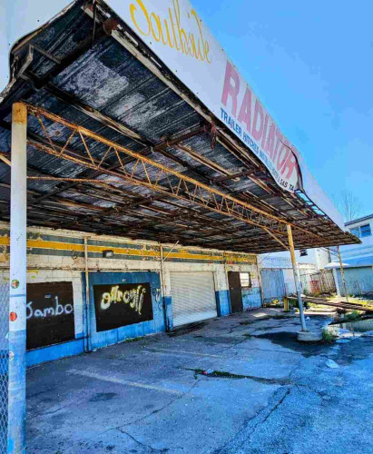 The side view, beneath the overhead canopy of an old, abandoned vehicle radiator repair shop, rusting and derelict. With dirt and debris, boarded up windows, graffiti, and cracked, peeling paint.
