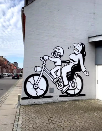 Streetartwall. A funny black and white mural with two cartoon characters and a bicycle was painted on the white exterior wall of a single-storey house. The mural is designed in the style of a caricature with simple, broad and clear lines. A little boy with a bicycle helmet is happily sitting on a bicycle and pedaling. His mother sits in the back of the child seat, slumped over and asleep. It has been a busy week. (In front of the mural is a paved forecourt and next to it a view of the street)
Artist: "HuskMitNavn" = Remember my name