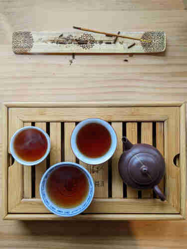 Three porcelain cups of various sizes filled with oolong tea next to a yixing teapot on a bamboo tray. Incense is burning on the table near the tea.