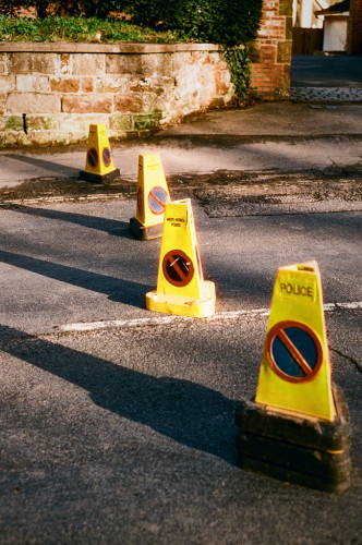 Portrait format colour photo showing four yellow, blue and red cones (the nearest simply labeled "POLICE", the next labeled "WEST MERCIA POLICE"), spaced across the road to close it. There is a stone wall in the background; the scene is lit by the afternoon sun, so the cones cast long diagonal shadows.