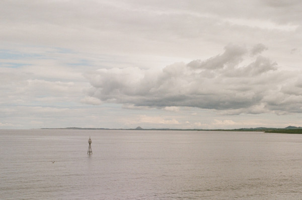 The bottom third of this colour photo shows a calm sea, with a marker post towards the left, and a seabird. Above is a thin strip of the coast of East Lothian with the cone of North Berwick Law visible. The top two thirds is sky mostly obscured by high clouds, very small patches of blue visible, and a larger low cloud to the right. Taken with a Pentax LX on Fuji Superia 400 film.