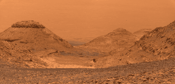 Panorama of Mars' Gale Crater, looking downhill from Mt. Sharp, colored in tones of red-orange. There are several smaller peaks downhill.