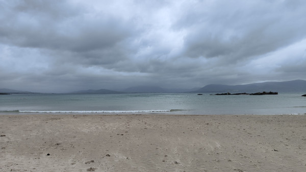 A beach on a day full of weather. The beach itself is fine beige same, and you can see patterns on the surface made by the wind. The sea is mostly flat apart from a small wave breaking towards the left hand  side, and it's a grey-green kind of colour. In the distance there are shadowy hills, which are partially concealed by mist and cloud. The sky is full of low, glowering grey clouds.