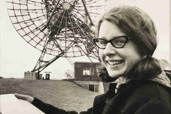 Photo of Jocelyn Bell Burnell in front of a radio telescope dish in the 1960s. She is a white woman with dark hair.