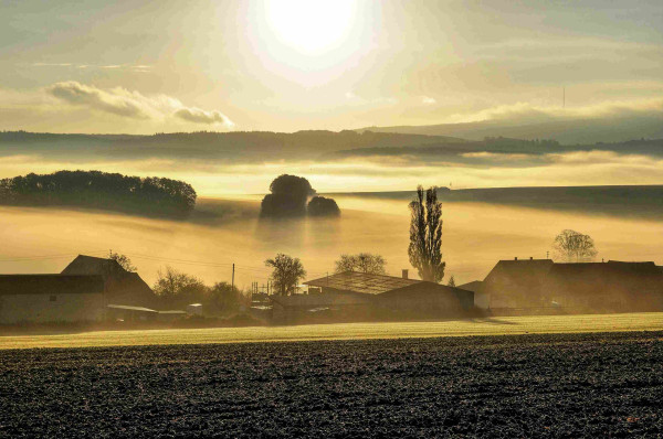 Landschaftsszenerie im Sonnenaufgang. Vorne eine Ackerfläche, dann eine kleine Ansiedlung und Hügellandschaft mit diffusen Nebelschwaden. 