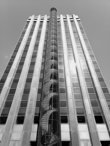 My photo with a black and white filter of a tall office building using the frog perspective so it looks wider at the bottom than the top with a spiral staircase in the middle 