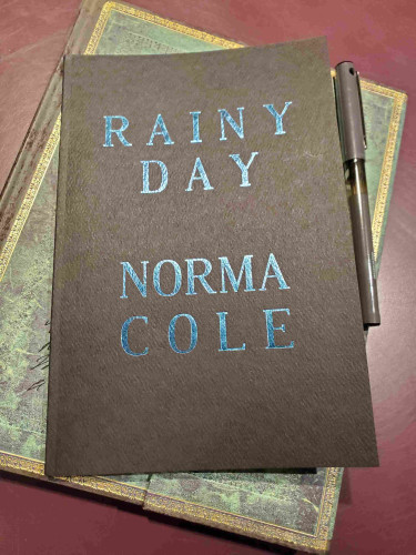 Poetry chapbook Rainy Day by Norma Cole (knife fork book), with its shiny blue lettering on a dark background, sits atop a notebook with an ornate green cover, with a black pen nearby