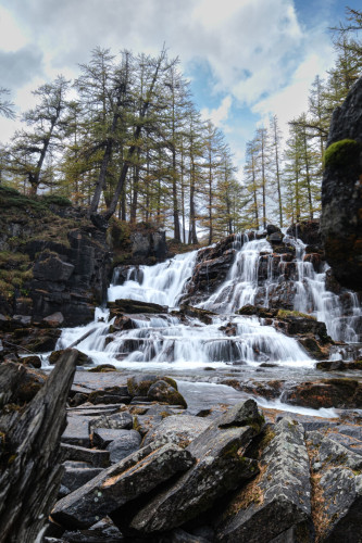 A cascading waterfall with multiple levels flowing over rocks, surrounded by pine trees under a partly cloudy sky.