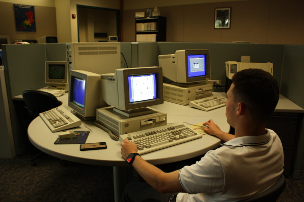 A twentysomething masculine-presenting person is playing SkiFree on a IBM PS/2, surrounded by other IBM PS/2s and a printer.