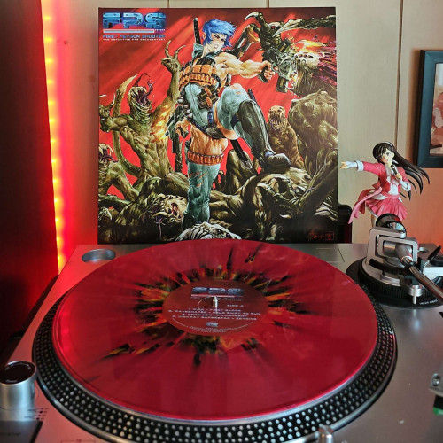 A Red w/ Black & Orange Splatter vinyl record sits on a turntable. Behind the turntable, a vinyl album outer sleeve is displayed. The front cover shows a woman fighting off alien monsters with a gun in each hand as she stands atop dead alien bodies. She is decked out in a leotard, tights, boots, and ammunition strapped around her.. 