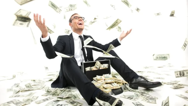 A man in a business suit, sitting on the floor with a suitcase full of money, throwing cash into the air and laughing.