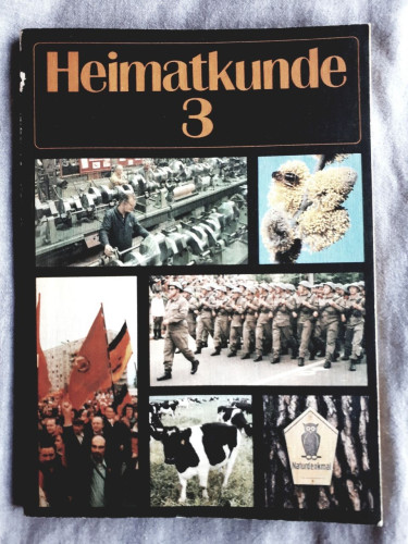 This book was used for primary school children in the GDR. After 1989, the topics of this type of social studies lesson changed. It is a black paperback book with photos. At the top is the title of the book, "Heimatkunde 3". The rest of the page is filled with 6 photos: in the left corner a picture of a factory with a man working on a machine; next to it a small close-up of flowers with a bee; in the middle a larger picture of a group of soldiers; in the left corner a photo of a demonstration with people carrying GDR flags; in the right corner a picture of a cow and a picture of the symbol for protected nature reservoir.
