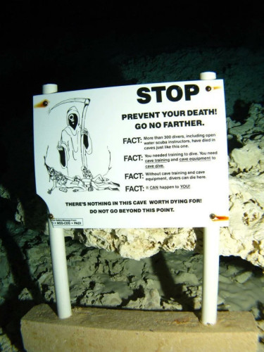 Sign on the ocean floor near some rock formations with a grim reaper standing over some skeletons. It reads ‘Stop. Prevent your death! Go no farther. FACT: More than 300 divers, including open water scuba instructors, have died in caves just like this one. FACT: You need training to cave dive. Without cave training and cave equipment, divers can die here.
FACT: It can happen to you! There’s nothing in this cave worth dying for!
Do not go behind this point.’