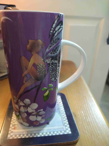 Tall mug of tea on a bedside cabinet, it is purple and has a Faerie sitting on white flowers staring at a crescent moon 
