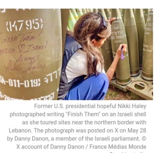 Haley kneeling and writing on a large military shell. The shell has the words "FINISH THEM! (America ♥ Israel) Always, Nikki Haley" written on it.