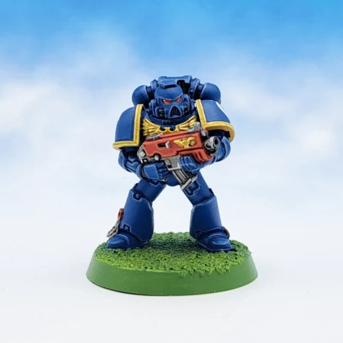 An Ultramarine Space Marine. He has blue armour, yellow trim on the shoulder pads, and a red bolt gun. The base has been painted in the classic "Goblin green" colour.