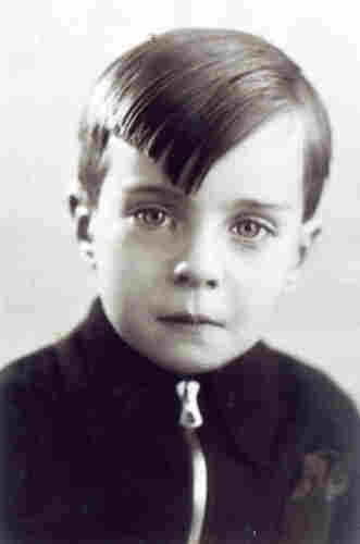 An ID portrait photo - face and shoulders - of a young boy. He has short hair but a long fringe falling over his right eye. She is wearing a zip-up sweatshirt - it is buttoned up to her neck. The boy has a slightly compressed mouth and very expressive, almost glazed eyes.