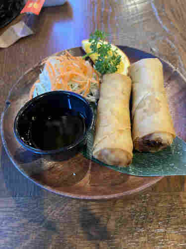 Plate with two spring rolls, dipping sauce, and a side of shredded vegetables, garnished with a lemon wedge and parsley, on a wooden table.