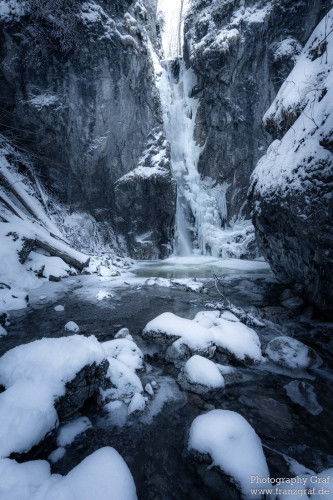 This is a stunning image of a natural landscape, presenting a striking contrast between the unyielding power of the waterfall and the serene stillness of the snowy mountain. The dominating force of the waterfall is seen rushing through the middle of the image, its icy waters tumbling down the rocky terrain. The surrounding area is blanketed in snow, creating an impression of tranquility and silence. The black shades in the image suggest it might be taken during the depth of winter. The scene seems to be of a remote, untouched location, possibly a hidden valley or ravine. The beauty of the image lies in its stark contrasts - the rush of the waterfall against the quiet of the snow, the harshness of the icy water against the softness of the snowfall, and the solitude of the wild landscape.