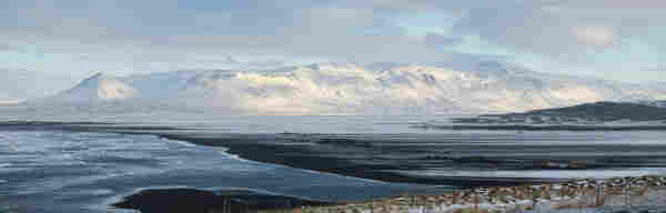 A wide panoramic photo of a beach scene. The foreground is scrubby brown grass covered in snow with fences running horizontally along it. Beyond that is a black beach with waves breaking from the left. Most of the sands look frozen. A river is running from the right and into the bottom centre of the shot to join the sea. On the horizon is a range of snow-covered mountains, lit from the right. Above them is a cloudy, pale cyan sky.