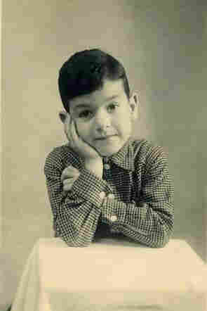 A young boy standing behind a small table. He is wearing a fine checked shirt. His hair is short. His fringe falls over half of his forehead. He rests his elbows on the table and supports his head on his right hand. He looks straight into the lens.
