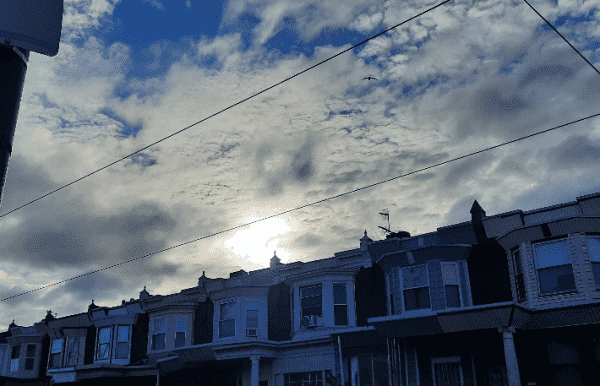 clouds and a blue sky above row homes. the sun shines brightly from behind the clouds. trolley lines cross the sky