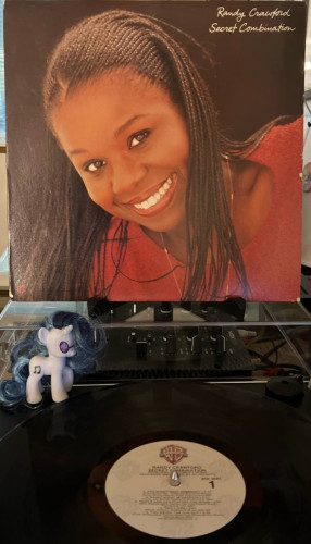 A record sleeve on a stand over a turntable. Sleeve has a picture of a smiling woman with French braids leaning into the frame. In the script in the top right corner "Randy Crawford" "Secret Combination"

On the turntable below is a record. The center label is beige with a Warner Bros. logo at the top.
