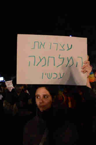 A woman holding a sign above her head that reads: “Stop the War Now” in Hebrew.