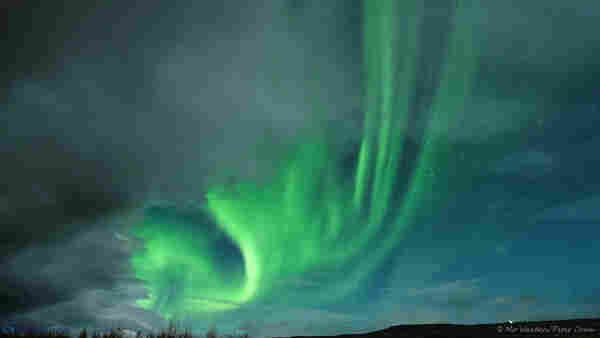 A photo of a night scene, almost completely filled by the sky. It's dark and some stars can be seen on the right, but a bank of heavy cloud is draped over the left side from top centre to bottom left. Peeping out from behind the cloud is a strong band of green aurora with feathery streaks climbing towards the top of the shot.
