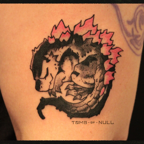 A fresh tattoo of a curled up, sleeping chibi Godzilla applied to olive skin. His expression is content like that of a snoozing cat.