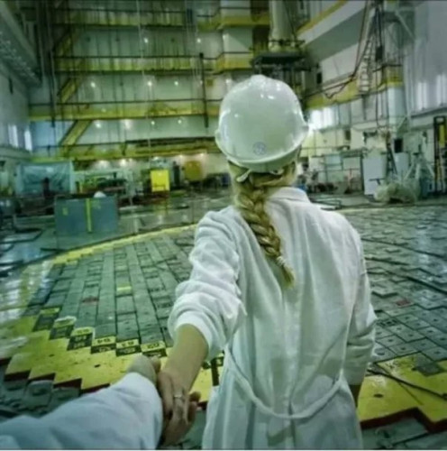 Still image. Person in an industrial white coat, white helmet, wearing long braided hair leads you by the hand down what appears to be the reactor hall of an RBMK power plant. The floor squares are located over the reactor's fuel channels. 