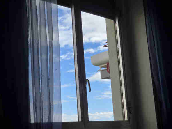 A serene view through an open window, framed by curtains, showcasing a slice of the tranquil blue sky with soft, white clouds drifting by. A white flower box is perched outside, hinting at the quiet domesticity within, as the scene bathes in the gentle glow of the afternoon sun. 