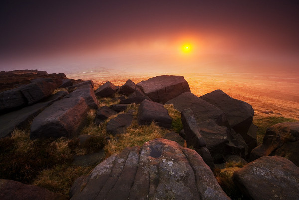 Photo of the sun rising into low cloud, casting a pink/red light on to a rocky outcrop in the foreground.